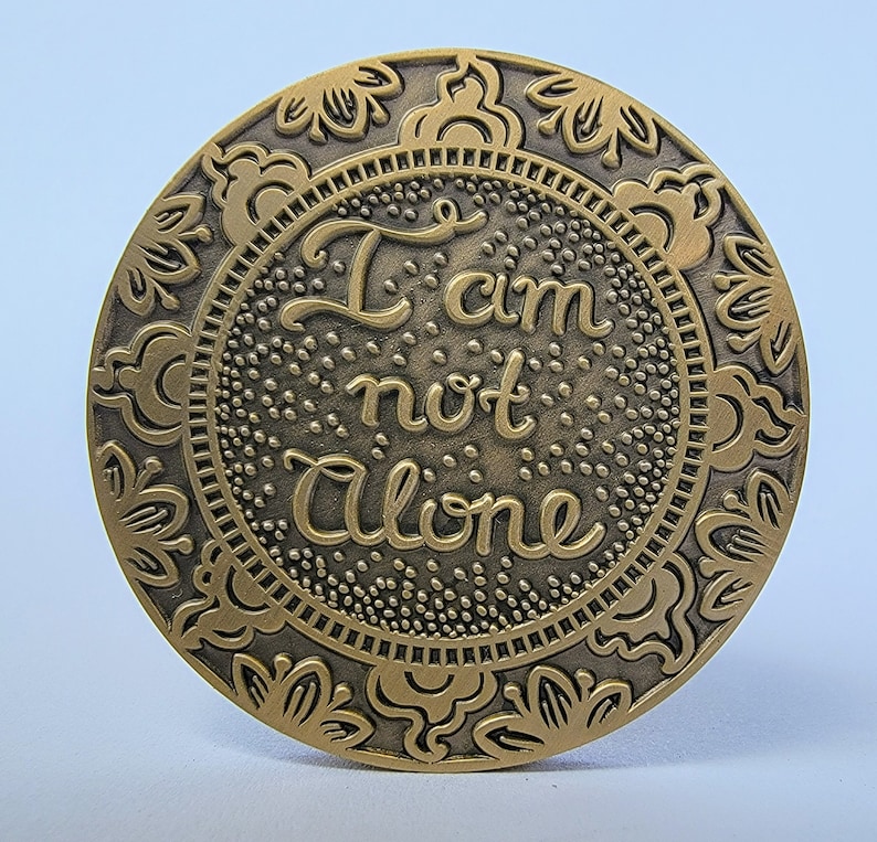 Pu&ograve; includere: Un gettone rotondo in metallo dorato con un design ornamentale. Il centro del gettone presenta le parole "Non sono solo" in un font stilizzato.