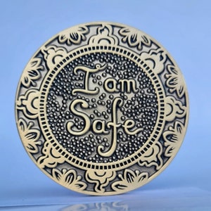 May include: A round metal token with an ornate border and the words "I am Safe" in a decorative font.