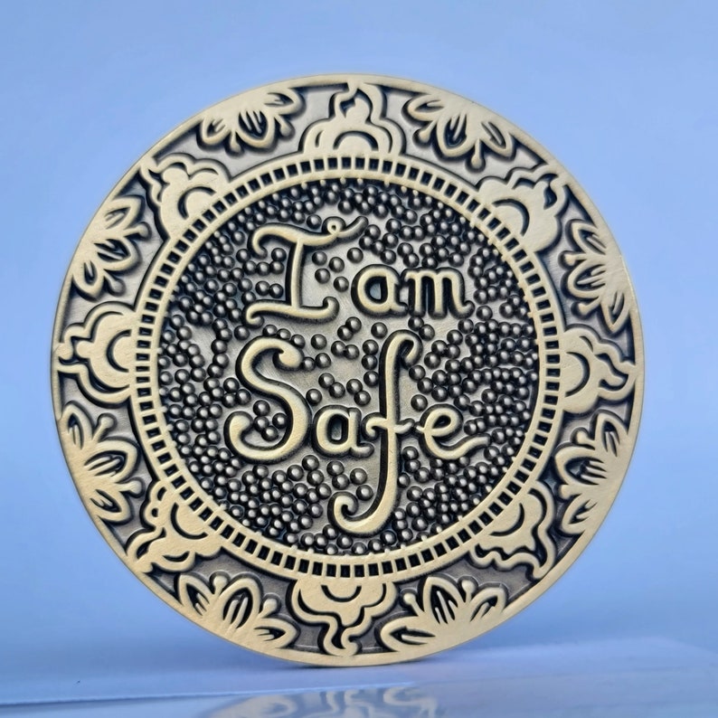 May include: A round metal token with an ornate border and the words "I am Safe" in a decorative font.