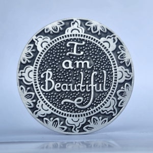 May include: A silver-toned round coin with an ornate floral design around the edge. The centre of the coin features the words "I am Beautiful" in a decorative script.