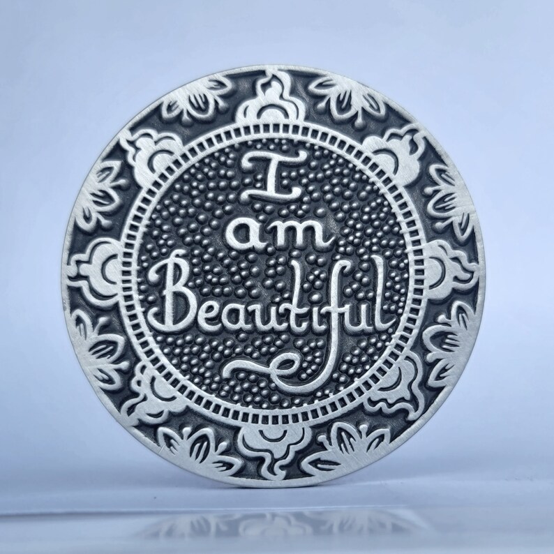 May include: A silver-toned round coin with an ornate floral design around the edge. The centre of the coin features the words "I am Beautiful" in a decorative script.