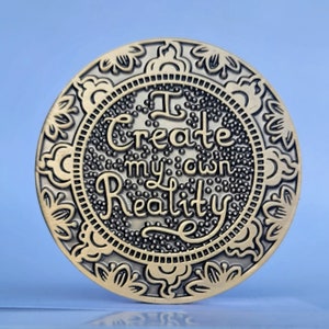 May include: A gold-toned round coin with an ornate border and the words "I create my own reality" in a decorative font.