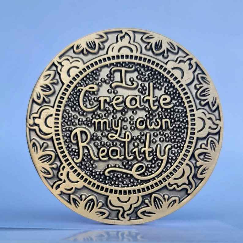 May include: A gold-toned round coin with an ornate border and the words "I create my own reality" in a decorative font.