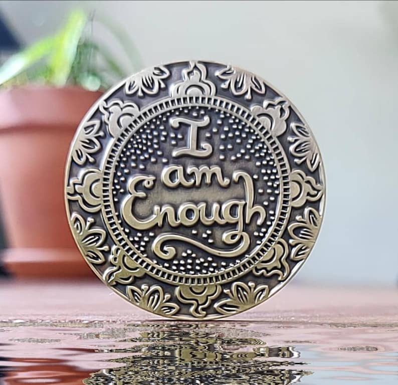 May include: A round, brass-coloured token with an ornate design and the words "I am Enough" inscribed in a decorative font.