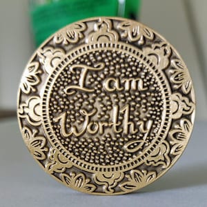 Pu&ograve; includere: Un gettone in metallo dorato con un design ornamentale attorno al bordo. Il centro del gettone presenta le parole "I am Worthy" in un carattere stilizzato.