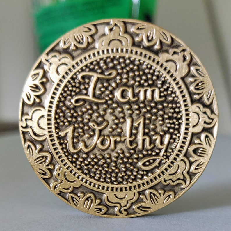 Pu&ograve; includere: Un gettone in metallo dorato con un design ornamentale attorno al bordo. Il centro del gettone presenta le parole "I am Worthy" in un carattere stilizzato.