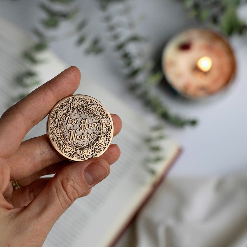 May include: A round, copper-coloured token with an intricate design and the words "Be Here Now" inscribed in a cursive font.