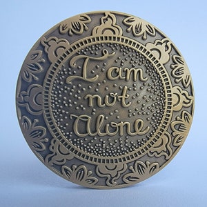 May include: A round, gold-coloured metal medallion with an ornate design around the edge. The centre of the medallion features the words "I am not alone" in a stylised font.