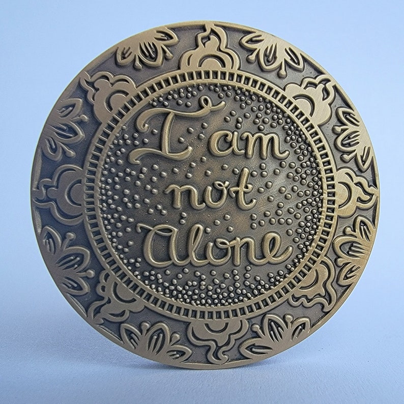 May include: A round, gold-coloured metal medallion with an ornate design around the edge. The centre of the medallion features the words "I am not alone" in a stylised font.
