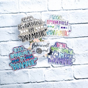 Könnte beinhalten: Vier glitzernde, bunte, runde, Plastikabzeichen mit dem Text "SOOO APPARENTLY... I'M "DRAMATIC"" auf jedem. Die Abzeichen befinden sich auf einem weißen Ziegelmauerhintergrund.