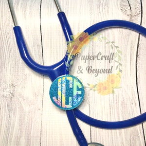 May include: A blue stethoscope with a round, glittery monogram charm. The charm features the initials "JCF" in a decorative font. The stethoscope is positioned on a light wood-grain surface. The text "PaperCraft & Beyond" is visible.
