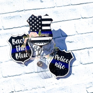 May include: Three black and white police badge-shaped resin ornaments with blue and white thin blue line accents. The ornaments have the text "Back the Blue", "Police Wife", and a US flag design.