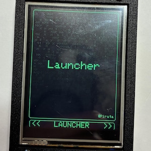 May include: A black rectangular electronic device with a screen displaying the green text Launcher and @Pirata. The screen features a green border and navigation arrows at the bottom. The device is secured with four silver screws in the corners.