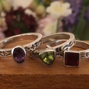 May include: Three silver rings with different colored gemstones. The first ring has a purple oval gemstone, the second ring has a green triangular gemstone, and the third ring has a red square gemstone. All rings have an intricate floral design.