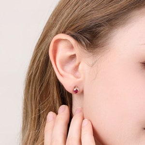 May include: A close-up of a woman's hand wearing a ring with a red gemstone. The ring is set in gold and has small diamonds on the band. The woman is also wearing a pair of earrings with red gemstones.