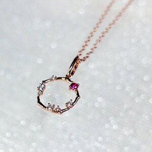 May include: A rose gold necklace featuring a delicate circular pendant with a textured, branch-like design. The pendant is accented with several small, clear white stones and one prominent, round, vibrant pink stone set along the thin metal ring.