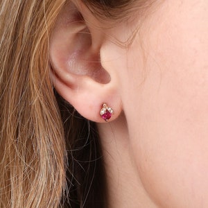 May include: A pair of small gold stud earrings with a single red gemstone and smaller white gemstones surrounding it.