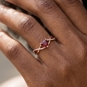 May include: A rose gold ring with a red gemstone in a square shape. The ring has a twisted band design with small, clear stones. The ring is on a person's finger.