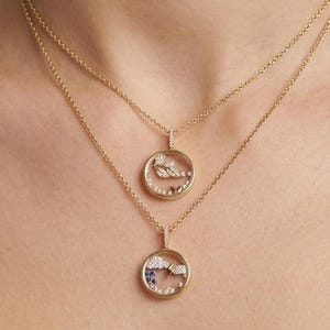 May include: Two gold necklaces with circular pendants. The pendants feature a design with small stones and a delicate frame. The necklaces are layered, with one slightly longer than the other. The background is blurred, with a soft, neutral color palette.