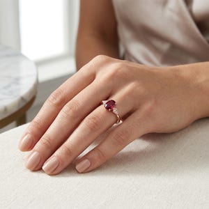May include: A rose gold ring featuring a large oval-cut ruby gemstone and two smaller triangular diamonds. The ring is set on a delicate band, showcasing the vibrant red of the ruby and the sparkle of the diamonds.