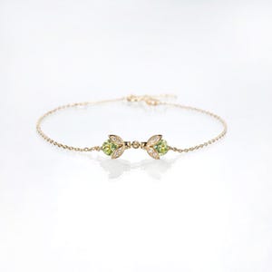 May include: A delicate gold-colored bracelet featuring a central design with two green gemstone clusters, each accented with small clear stones. The bracelet has a fine chain and a clasp.