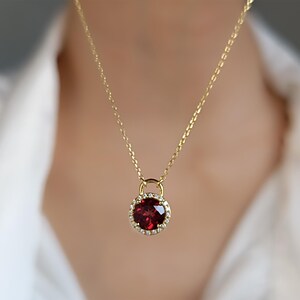 May include: A gold chain necklace with a pendant featuring a round red gemstone surrounded by a halo of smaller white gemstones.