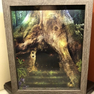 May include: Framed art print depicting a fantasy scene of a tree with a cave entrance. The artwork features a butterfly, lanterns, and a mystical forest setting. The frame is a light gray color.
