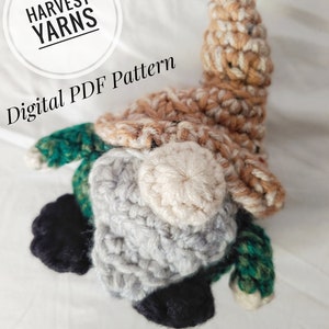 May include: A crocheted gnome in shades of brown, beige, grey, green and black. The image includes the text "HARVEST YARNS" and "Digital PDF Pattern".