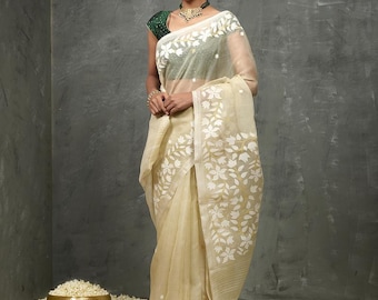 May include: A woman wearing a cream-colored saree with white floral embroidery. The saree is sheer and has a delicate, flowing drape. The woman is standing on a bed of white flowers.