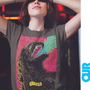 May include: Dark gray t-shirt featuring a Godzilla graphic. The graphic shows Godzilla roaring with a blue and yellow energy blast against a pink background. The word "GODZILLA" is printed in yellow at the bottom of the graphic.