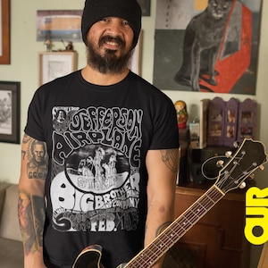 May include: A man wearing a black t-shirt with a white graphic of a concert poster for Jefferson Airplane. The poster features the band's name, the date "Feb. 10, '68", and the words "Big Brother and the Holding Company". The man is also wearing a black beanie and has a beard. He is holding a brown and black electric guitar.