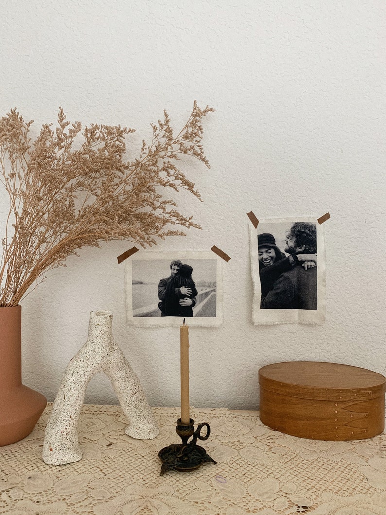 Puede incluir: Dos fotograf&iacute;as enmarcadas en blanco y negro de parejas abraz&aacute;ndose, expuestas en una pared blanca con un jarr&oacute;n marr&oacute;n de flores secas y un jarr&oacute;n de cer&aacute;mica blanca. Un candelero de lat&oacute;n con una vela encendida se encuentra sobre un mantel de encaje blanco con una caja de madera.
