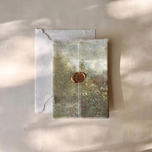 Puede incluir: Una tarjeta rectangular con una impresión de un paisaje impresionista de un jardín con árboles y flores, asegurada con un sello de cera dorado metálico circular, colocada sobre un sobre blanco liso con un acabado de borde irregular.