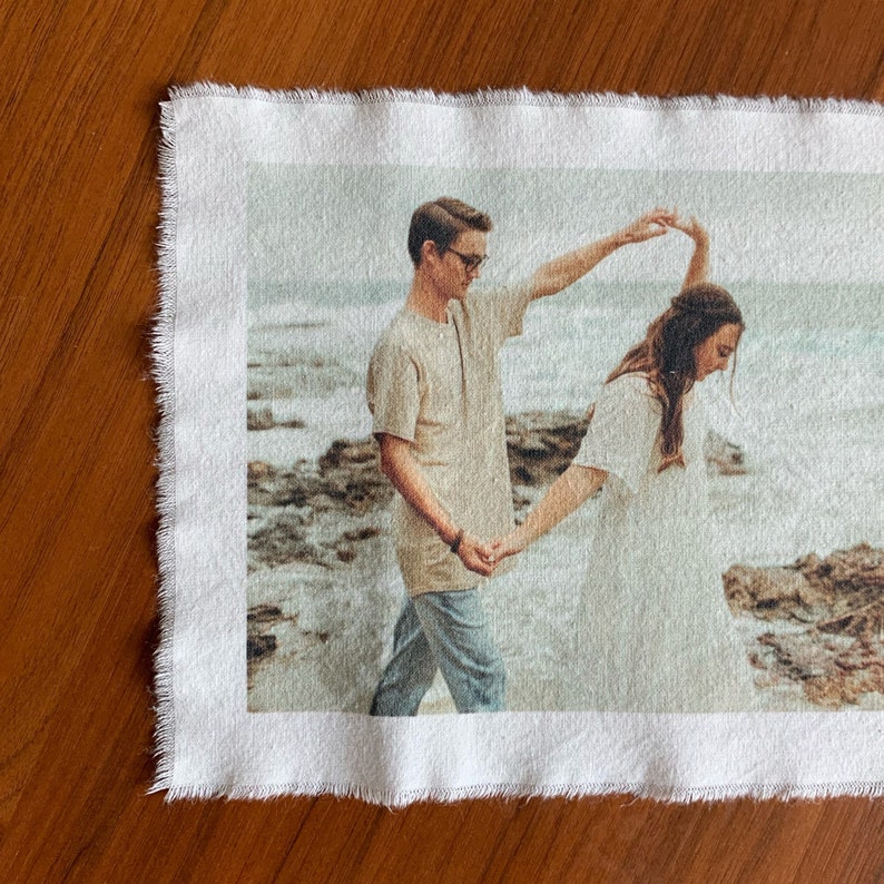Puede incluir: Una pareja se toma de la mano y gira en una playa. El hombre lleva una camisa beige y pantalones vaqueros azules. La mujer lleva un vestido blanco. El fondo es una playa con rocas y agua.