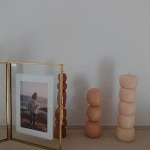 Puede incluir: Una foto enmarcada en oro con una foto de un ni&ntilde;o con un sombrero de pie en una playa. Tres velas redondas apiladas en tonos marrones y beige est&aacute;n en un estante al lado de la foto.