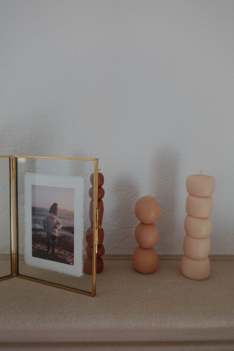 Puede incluir: Una foto enmarcada en oro con una foto de un ni&ntilde;o con un sombrero de pie en una playa. Tres velas redondas apiladas en tonos marrones y beige est&aacute;n en un estante al lado de la foto.
