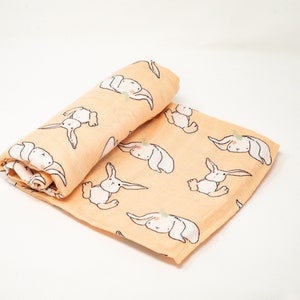 May include: A peach-colored muslin swaddle blanket with a repeating pattern of white bunnies. The bunnies are drawn in a simple, whimsical style.