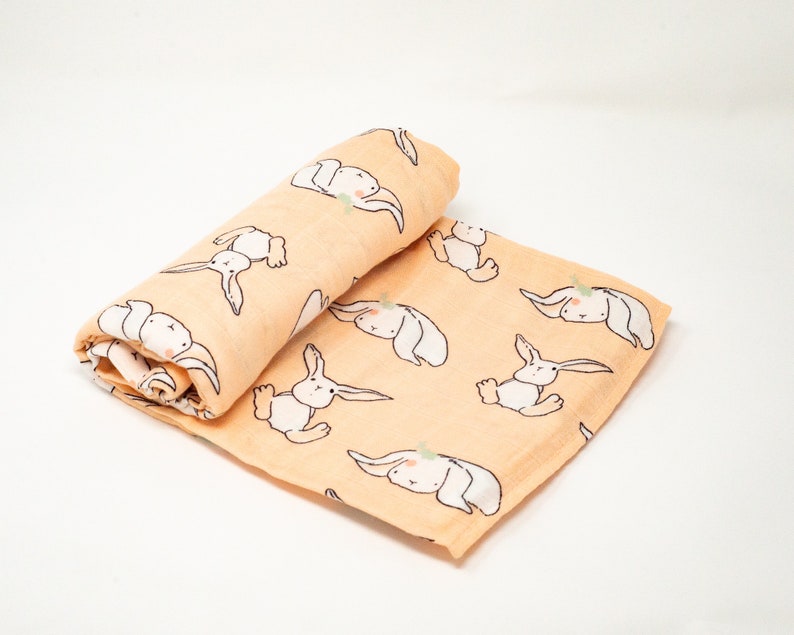 May include: A peach-colored muslin swaddle blanket with a repeating pattern of white bunnies. The bunnies are drawn in a simple, whimsical style.