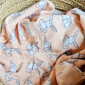 May include: A peach-colored muslin baby blanket with a white bunny rabbit pattern. The blanket is folded and placed in a woven basket.