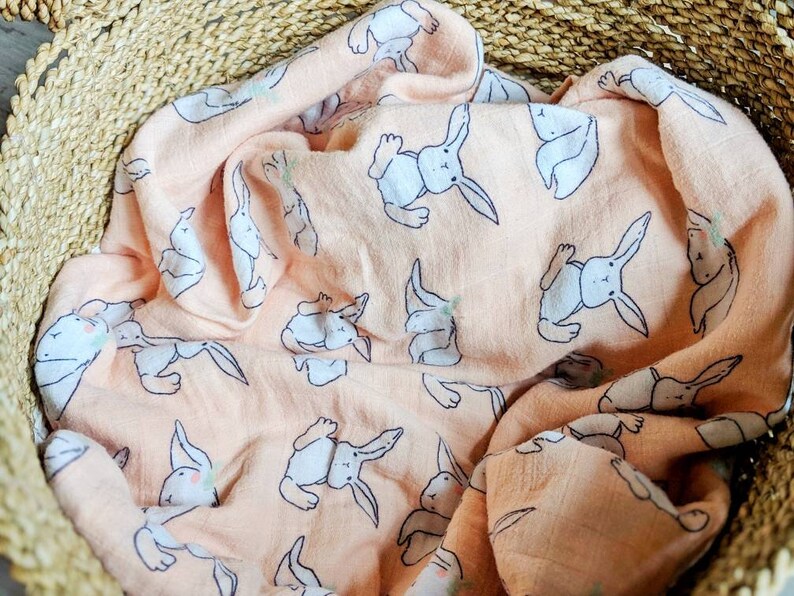 May include: A peach-colored muslin baby blanket with a white bunny rabbit pattern. The blanket is folded and placed in a woven basket.