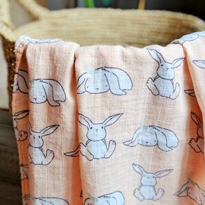 May include: A peach-colored muslin swaddle blanket with a repeating pattern of gray bunnies. The bunnies have small flowers on their heads and are in various poses.