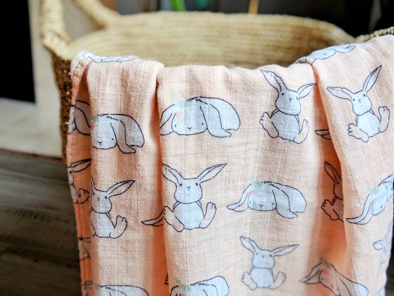 May include: A peach-colored muslin swaddle blanket with a repeating pattern of gray bunnies. The bunnies have small flowers on their heads and are in various poses.