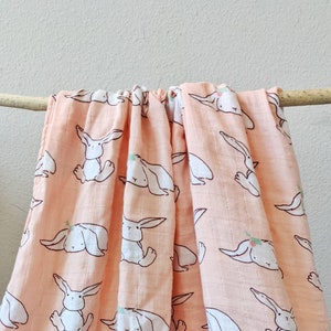 May include: A peach-colored muslin baby blanket with a repeating pattern of white bunnies with floral accents. The blanket is folded and draped over a branch.