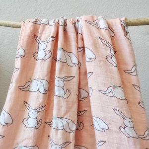 May include: A peach-colored muslin baby blanket with a repeating pattern of white bunnies wearing little crowns. The blanket is made of soft, breathable fabric and is perfect for swaddling or cuddling.