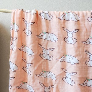 May include: A peach-colored muslin baby blanket with a repeating pattern of white bunnies with green floral headbands.