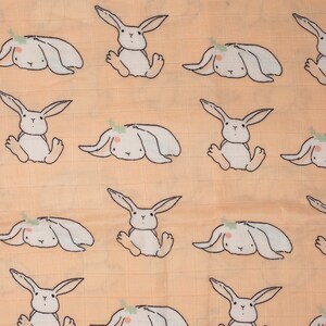 May include: A peach-colored fabric with a repeating pattern of white bunnies. The bunnies have small, green leaves on their heads and are in various poses.