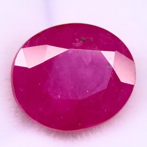 Puede incluir: Una gema de rubí facetada, de forma ovalada. La gema es de un color rojo intenso y saturado con una superficie pulida. Las facetas crean un juego de luces y sombras.