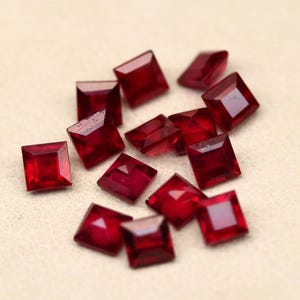 May include: A collection of square-cut, deep red gemstones. The gems are scattered on a light beige surface, each displaying facets that catch the light. The stones appear to be approximately 0.25 inches in size, with sharp, defined edges.