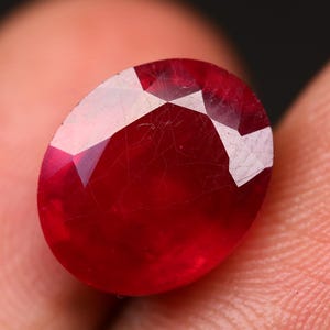 Könnte beinhalten: Nahaufnahme eines facettierten, oval geformten Rubin-Edelsteins. Der Edelstein hat eine tiefrote Farbe mit weißen Facetten, gehalten zwischen Fingern. Die Oberfläche des Rubins weist sichtbare Einschlüsse und Unvollkommenheiten auf.