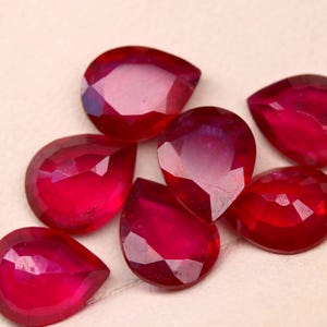 May include: Seven faceted, teardrop-shaped rubies are displayed. The gemstones are a deep red colour, with a high degree of clarity, reflecting light. The rubies are arranged on a light-coloured surface.